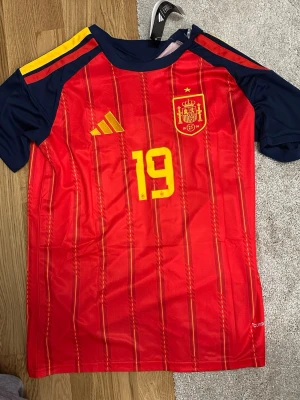 Spaniens matchtröja Lamine Yamal #19 - Röd och marinblå matchtröja från Adidas för Spaniens landslag med gula detaljer, tryckt nummer 19 och Lamine Yamal på ryggen. Tröjan har korta ärmar, tunna vertikala ränder och broderad landslagssköld på bröstet. Tillverkad i lätt funktionsmaterial.