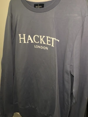 Blå sweatshirt från Hackett London - Snygg blå sweatshirt från Hackett London med vit logga tryckt på bröstet. Tröjan har rund halsringning och ribbade muddar vid ärmslut och nederkant. Perfekt för en avslappnad och stilren look.