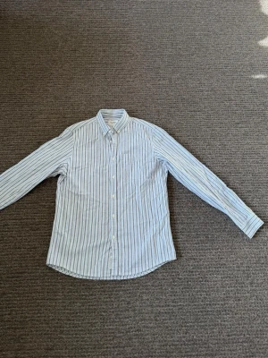 Randig skjorta  - Klassisk skjorta från pull and bear med vita och blå vertikala ränder. Skjortan har lång ärm, knappar framtill och en tidlös krage. Tillverkad i bomull för en skön och luftig känsla. Perfekt för en clean och stilren look.