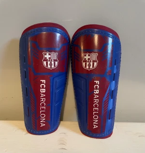 Blå-röda benskydd FC Barcelona - Säljer ett par blå och röda benskydd från FC Barcelona med klubbens logga och text på framsidan. Skydden har en hård plastyta och mjuk vaddering på insidan för extra komfort. Perfekta för fotbollsträning eller match. 21,5 cm