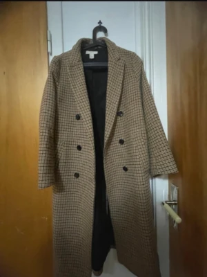 Rutig brun lång rock kappa med dubbelknäppning - Snygg lång rock i brunt med svart rutmönster och dubbelknäppning framtill. Klassisk krage och rak passform, helfodrad med svart innerfoder. Perfekt för dig som gillar tidlös och trendig stil. Märke och material framgår ej.