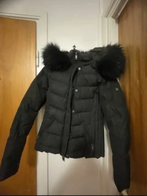 Svart pufferjacka med fuskpäls - Snygg svart pufferjacka med stor huva och fluffig fuskpäls runt kanten. Jackan har dragkedja och knappar framtill, två snedställda fickor och markerad midja för en schysst siluett. Perfekt för kalla vinterdagar och riktigt varm.