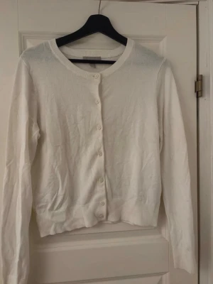 Vit basic kofta med rund hals - En enkel och stilren vit kofta med rund halsringning och knäppning framtill. Långärmad modell med ribbade muddar vid ärmslut och nederkant. 