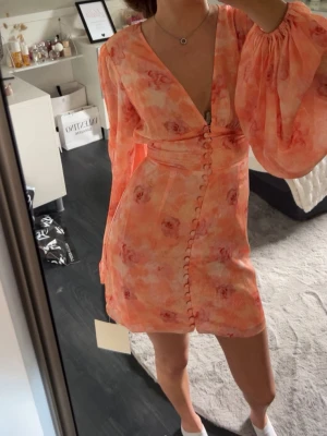 Blommig kortklänning med puffärm - Säljer en superfin kort klänning i ljusa orange och rosa toner med blommigt mönster. Klänningen har djup v-ringning, puffiga långärmar och knappar framtill. Markerad midja och lätt volang nedtill ger en feminin siluett. Perfekt för sommarens alla tillfällen!