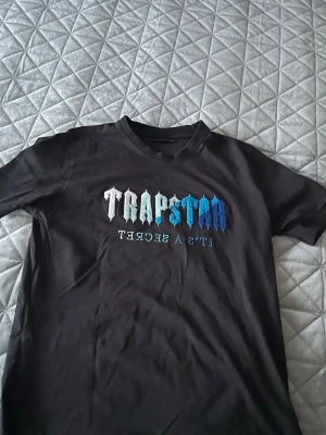 Trapstar svart t-shirt med blå text - Säljer en svart t-shirt från Trapstar med broderad text i vitt och blått på bröstet där det står 'Trapstar It's a secret'. T-shirten har klassisk passform och korta ärmar. Perfekt för dig som gillar streetwear och vill sticka ut.