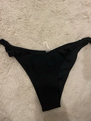 Svart bikiniunderdel med volang - Snygg svart bikiniunderdel med volangdetalj i midjan och klassisk trosa-modell. Tillverkad i mjukt och stretchigt material som passar perfekt till sommaren. Enkel och stilren design som är lätt att matcha med olika överdelar. aldrig använt 