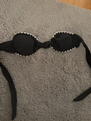 Svart bikiniöverdel med vita detaljer - Snygg svart bikiniöverdel med vita sicksack-detaljer längs kanten. Bandeau-modell med knyt i ryggen och framsidan. Aldrig använd, ny utan prislapp 