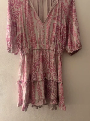 Rosa och vit kortklänning med volang - Söt kortklänning i rosa och vitt med blommigt mönster och volangdetaljer. Klänningen har v-ringning, resår i midjan och korta puffärmar. Perfekt för sommarens alla tillfällen.