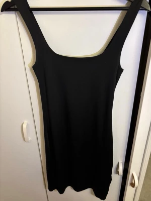 Svart klänning från Amisu - En enkel och stilren svart klänning från Amisu. Klänningen är ärmlös med breda axelband och har en rak, kroppsnära passform. Tillverkad i mjukt material och ser satin aktig ut.