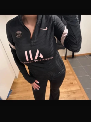 Svart/Rosa PSG träningströja från Nike - Snygg svart långärmad träningströja från Nike x PSG med halv dragkedja, rosa detaljer och PSG-logga på bröstet. Perfekt för träning eller sportiga dagar.