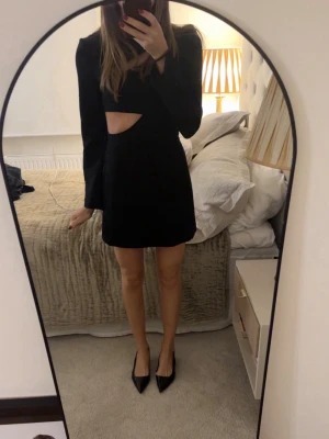 Svart kort klänning med cut out - Trendig svart kort klänning med lång ärm och snygg cut out-detalj vid midjan. Klänningen har en figurnära passform och är perfekt för dig som vill ha en stilren men edgy look. Materialet känns mjukt och följsamt.