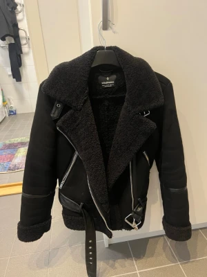 Svart teddyjacka från Stradivarius - Snygg svart teddyjacka från Stradivarius med bred krage, dragkedjor och bälte i midjan. Jackan har mjukt teddyfoder, detaljer i fuskskinn och muddar vid ärmsluten. Perfekt för kalla dagar och ger en cool look.