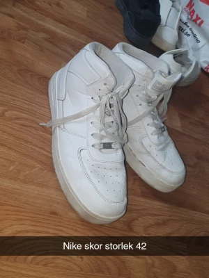 Vita Nike high top sneakers - Säljer ett par klassiska vita high top sneakers från Nike. Skorna har en stilren design med perforerade detaljer på tån, vadderad krage och snörning framtill. Tillverkade i läder med textilfoder för extra komfort. Perfekta för en clean och sportig look.