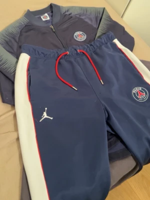 PSG x Jordan blå träningsset - Snyggt träningsset från PSG x Jordan med jacka och byxor i marinblått med vita och röda detaljer. Jackan har dragkedja och PSG-logga på bröstet, byxorna har resårmidja med röda snören, vita sidopaneler och Jordan-logga. Perfekt för fotboll eller chill.