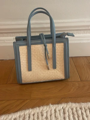 Blå och beige handväska i rottingstil - Snygg handväska med ljusblåa kanter och handtag samt beige flätad rottingliknande mittparti. Väskan har en boxig form och dubbla handtag, perfekt för dig som gillar trendiga accessoarer med en touch av sommar. Stängs med dragkedja och har dekorativa detaljer i blått.