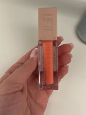 Maybelline Lifter Gloss Peach Ring - Maybelline Lifter Gloss i färgen 22 Peach Ring. Ett läppglans med persikotonad nyans och glossig finish. Kommer i en stilren, genomskinlig plastförpackning med beige lock och stor applikator. Innehåller hyaluronsyra för extra återfuktning.