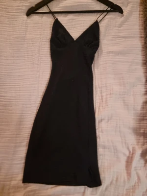 Svart långklänning från ICI COI - Svart långklänning med smala axelband som korsas i ryggen. Klänningen har v-ringad urringning och är figurnära med en liten slits nedtill. Materialet är strukturerat och känns mjukt mot huden. Perfekt för dig som gillar stilrena och enkla plagg.