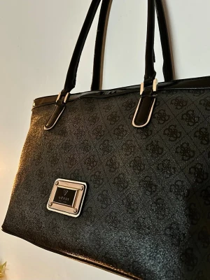 Guess Tote Bag "Coal" – Perfekt skoväska/laptopväska! ✨💻 - Säljer min rymliga Guess-tote i klassiskt monogram-mönster! 🖤 Perfekt för skolan/jobbet då den rymmer ALLT. ✨ Mått: Bredd 44cm (topp) / 36cm (botten). Höjd inkl handtag ca 50cm. Rymmer enkelt laptop & mappar! ✨ Skick: Använt skick Väskan är använd och har lite kärleksskav, Insidan & fodret är superfräscht! Säljes därför till ett grymt fyndpris! ✨ Nypris: Ca 1800kr.