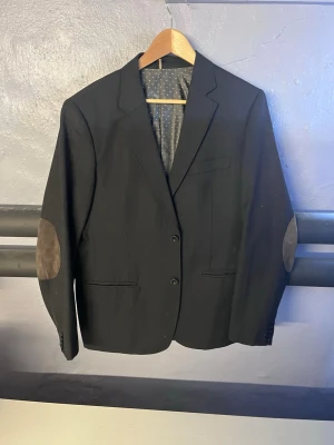 Svart kavaj från L'Ecole - Stilren svart kavaj från L'Ecole med två knappar framtill och bruna mockadetaljer på armbågarna. Fodret är grått med vita prickar för en cool kontrast. Perfekt för dig som vill ha en klassisk men ändå trendig look.