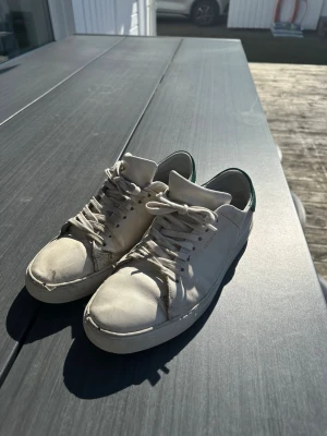 Vita sneakers från Axel Arigato clean 90 sneaker  - Stilrena vita sneakers från Axel Arigato med lågt skaft och klassisk siluett. Skolåda från Axel arigato följer med nyskick Skorna har vita snören, diskret logga på sidan och grön detalj vid hälen. Tillverkade i läder för en clean look som passar till det mesta.