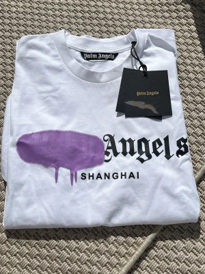 Vit Palm Angels t-shirt Shanghai - Säljer en vit t-shirt från Palm Angels med trycket 'Shanghai' och ett lila grafiskt motiv över bröstet. T-shirten har korta ärmar, rund halsringning och är gjord i mjuk bomull. Perfekt för dig som gillar streetwear och vill sticka ut.