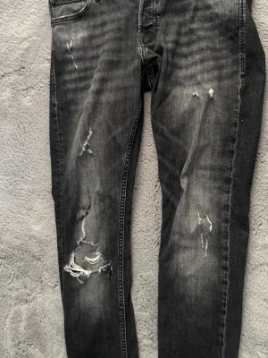 Svarta jeans Jack and Jones skinny  - Säljer ett par svarta jeans med slitningar och hål på benen. Jeansen har en klassisk femficksmodell och är tillverkade i denim med stretch. Perfekta för dig som gillar en edgy och trendig stil.