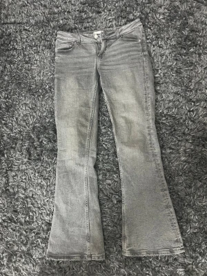 Grå bootcut jeans från Gina Tricot - Säljer ett par gråa bootcut jeans från Gina Tricot med klassisk femficksmodell och knappgylf. Jeansen har en snygg tvättad look och utsvängda ben som ger en retrovibe. Perfekta för dig som gillar en avslappnad men trendig stil.