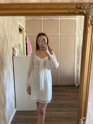 Vit kort klänning från NA-KD - Säljer en vit kort klänning från NA-KD med långa transparenta ärmar, v-ringning och knytband framtill. Klänningen har en feminin passform och är tillverkad i ett lätt material som ger en luftig känsla. Perfekt inför studenten!