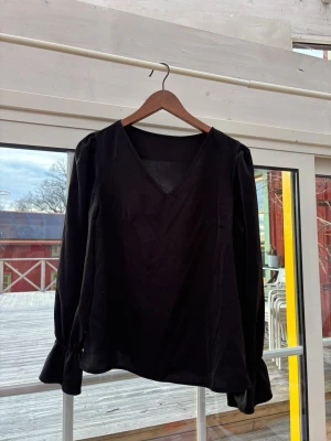 Svart blus med v-ringning - Stilren svart blus med v-ringad hals och långa ärmar. Ärmarna har en snygg rynkning vid mudden som ger en trendig touch. Tillverkad i ett mjukt, lätt material som faller fint. Perfekt att matcha med jeans eller kjol för en cool look.