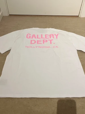 Vit Gallery Dept. t-shirt med rosa tryck - Vit t-shirt från Gallery Dept. med rosa texttryck både fram och bak. På ryggen står det 'GALLERY DEPT. HOLLYWOOD, CA.' i stor rosa text. Klassisk passform och rund hals. Perfekt för dig som gillar streetwear och tydliga loggorna. Sitter baggy