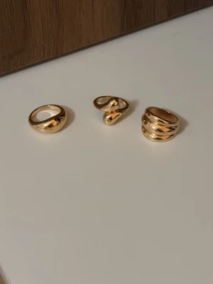 Guldfärgade ringar från Montini - Säljer ett set med tre guldfärgade ringar från Montini. Ringarna har olika former: en klassisk rund, en med tvinnad design och en med öppen, vågig form. Tillverkade av oädla material och perfekta för att ge din look en trendig touch. De är antingen i storlek 17,18 eller 19 är ej säker. Har aldrig använt