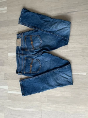 Blå jeans från Nudie Jeans - Säljer ett par klassiska blå jeans från Nudie Jeans med snygga kontrastsömmar på bakfickorna och normal passform. Jeansen har fem fickor och är gjorda i slitstarkt denimtyg. Perfekta till en avslappnad stil.