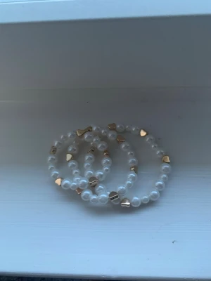 Pärlarmband med guldfärgade hjärtan - Säljer ett snyggt armband med vita pärlor och små guldfärgade hjärtan. Armbandet har en elastisk tråd som gör det enkelt att ta på och av. Perfekt accessoar för att lyfta din outfit med en söt och trendig touch.