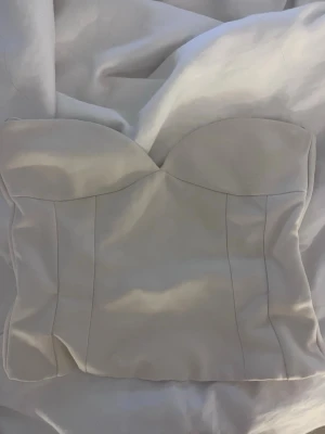 Vit axelbandslös korsettopp - Säljer en stilren vit korsettopp med sweetheart-urringning och markerade sömmar. Toppen är axelbandslös och har dragkedja i ryggen för enkel påklädning. Perfekt för att skapa en trendig och elegant look.