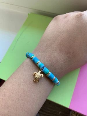 Turkost pärlarmband med sköldpaddshänge - Snyggt armband med turkosa och guldiga platta pärlor samt ett guldfärgat sköldpaddshänge. Armbandet har en fräsch och färgglad look som sticker ut och passar perfekt för dig som gillar detaljer. Elastiskt band för enkel påtagning.