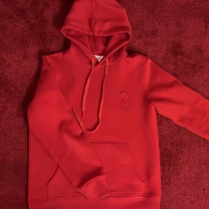 Röd AMI Paris hoodie - Snygg röd hoodie med huva och dragsnören. Klassisk känguruficka framtill och diskret broderad detalj på bröstet. Perfekt för en avslappnad och trendig look. Tillverkad i mjukt material som känns skönt mot huden. 