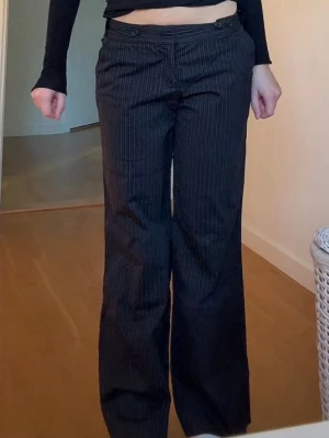 Svarta kostymbyxor med smal rand - Säljer ett par svarta kostymbyxor med diskreta vita nålrandiga detaljer. Byxorna har en rak, något utsvängd passform och stängs med knapp och dragkedja framtill. Perfekta för en stilren och trendig look.