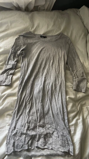 Grå t-shirt klänning med spets från Fransa - Mysigt klänning från Fransa i mjukt material. Modellen har trekvartsärmar och rund halsringning. Nedtill finns en bred spetskant som ger en söt och feminin touch. Perfekt för chillkvällar hemma