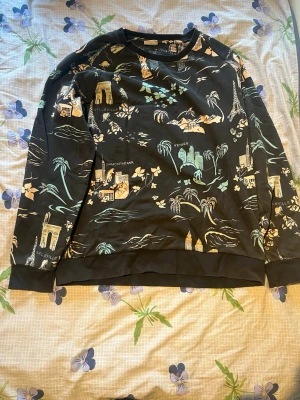 Svart hoodie med stadsmotiv - Cool svart hoodie med tryck av olika stadsmotiv som Eiffeltornet och palmer i beige, grönt och vitt. Mjuk bomullskänsla och avslappnad passform med ribbade muddar vid ärmar och midja. Perfekt för dig som gillar unika prints och streetstyle.