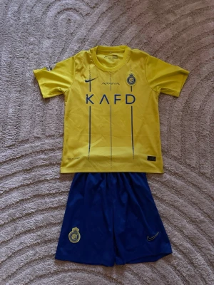 Al Nassr Ronaldo matchställ Nike - Säljer ett gult och blått Al Nassr matchställ med Ronaldo 7-tryck på ryggen. Tröjan har klubbmärke och sponsorloggor framtill, samt korta ärmar. Shortsen är blå med klubbmärke och Nike-logga. Perfekt för fotbollsträning eller att bära som supporter.