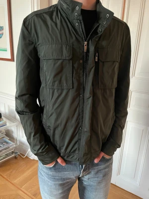 Mörkgrön bomberjacka med fickor - Snygg mörkgrön bomberjacka med hög krage och dragkedja framtill. Jackan har två stora bröstfickor med lock och sidofickor, samt detaljerade sömmar på axlarna. Perfekt för dig som vill ha en stilren och funktionell jacka. Jackan passar både M och S.