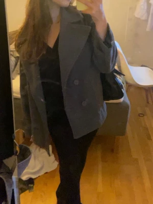 Grå kappa från Design by Si - Grå kappa i ullmix från Design by Si med bred krage och dubbelknäppning. Rak modell med helsvart foder som ger en clean look. Perfekt för höst och vinter när du vill vara både varm och snygg.