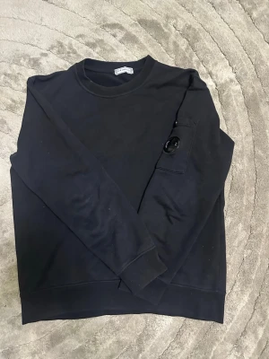 Svart långärmad topp från C.P. Company - Svart långärmad topp från C.P. Company med rund hals och en cool detalj på ärmen i form av en ficka med glaslins. Tillverkad i mjuk bomull och har en normal passning. Perfekt för dig som gillar stilrena plagg med unik touch.