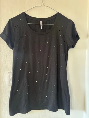 Svart t-shirt med pärldetaljer - Snygg svart t-shirt med korta ärmar och rund halsringning. T-shirten är dekorerad med små pärlor och diskreta håldetaljer framtill som ger en cool och trendig look. Perfekt att matcha med jeans eller kjol för en avslappnad stil.