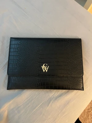 Svart datorväska  - Snygg svart datorväska i crocopräglat konstläder från CW. Väskan har ett stilrent kuvertformat lock och guldfärgad logga på framsidan. Perfekt för att bära din laptop med stil och hålla den skyddad i väskan.