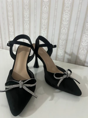 Svarta pumps med strassrosett - Snygga svarta pumps med spetsig tå och dekorativ strassrosett framtill. Skorna har en elegant rem runt ankeln och en unik, böjd klack. Materialet ser ut som mocka och de har en feminin och trendig look.