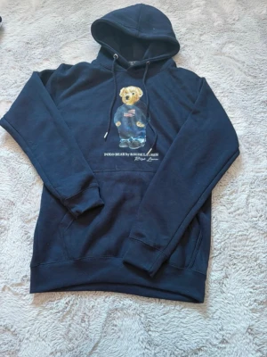 Mörkblå Polo Bear hoodie Ralph Lauren - Snygg mörkblå hoodie från Ralph Lauren med den ikoniska Polo Bear-trycket på bröstet. Hoodien har huva med dragsko och en stor magficka. Tillverkad i mjukt bomullsmaterial som är perfekt för chill dagar. Klassisk och stilren design som alltid funkar. Ny utan prislapp