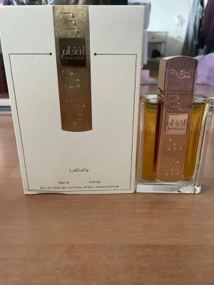 Lattafa Angham Eau de Parfum 100ml - Lattafa Angham Eau de Parfum kommer i en elegant, rektangulär glasflaska med guldfärgad metallkork och detaljer. Doften är 100 ml och förpackningen är vit med guldfärgade inslag och arabiska inskriptioner. Tillverkad i Förenade Arabemiraten.