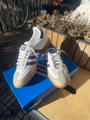 Adidas Samba OG vita/lila sneakers - Adidas Samba OG sneakers i vitt och lila med klassiska tre ränder på sidan. Ovandelen är i läder och mocka, med gummisula i brunt. Ikonisk retrostil som passar till streetwear och casual outfits. Skön passform och snygg design.