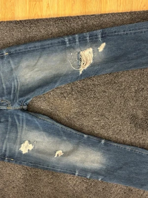 Blå slitna jeans med hål - Säljer ett par blå jeans med slitningar och hål på både lår och knä. Jeansen har en klassisk femficksmodell och är i ett mjukt denimtyg. Passformen är smal och tvätten är ljusblå med snygga slitningar för en trendig look. Priset går att diskuteras vid snabb affär